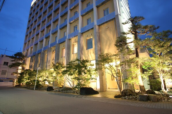 A multi-story building with a modern architectural design, featuring a series of windows and a well-maintained garden with trees and rocks. - Uozu Manten Hotel Ekimae (Uozu)