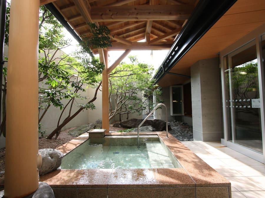 A tranquil outdoor pool area with a wooden pergola, stone seating, and a glass door leading to a building.