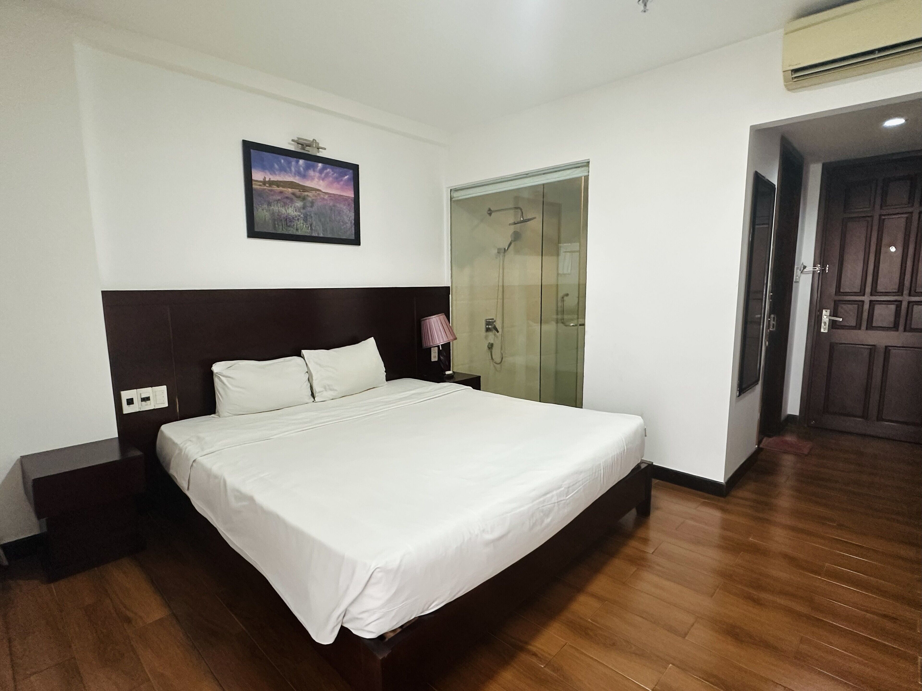 A hotel room with a large bed, a nightstand, a bedside lamp, a glass-enclosed shower, and a door to another room.