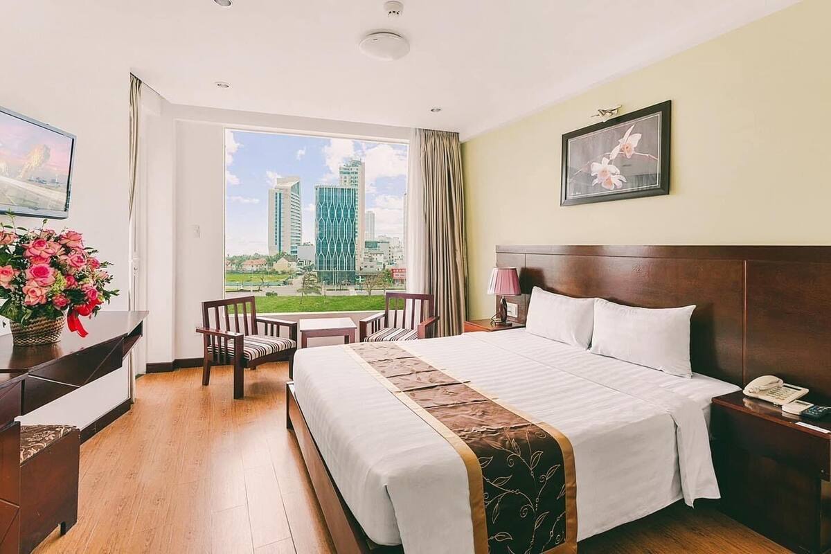 A hotel room with a large bed, a desk with a chair, a small table, a view of the cityscape, and a painting on the wall.