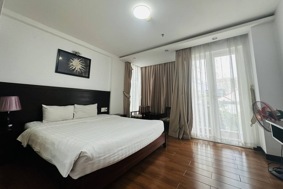 A hotel room with a large bed, a headboard, a nightstand, a lamp, a fan, and a window with curtains.