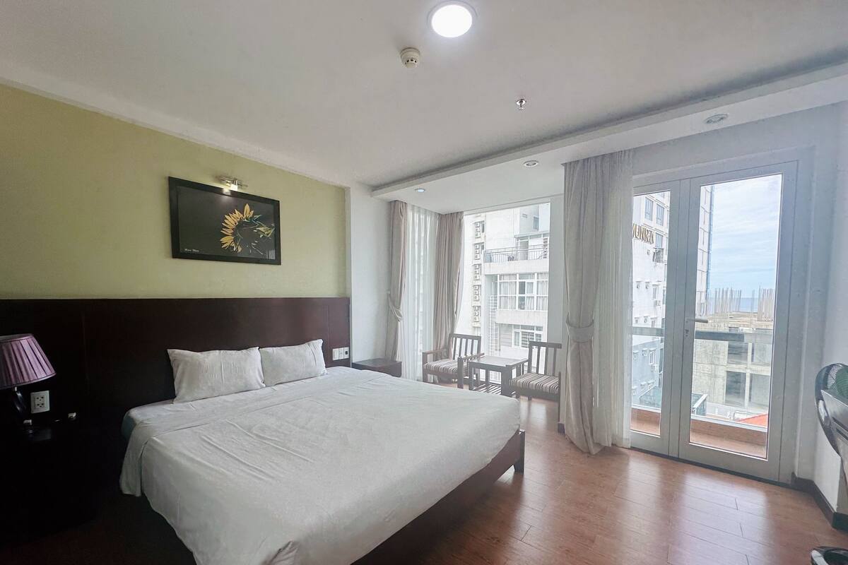 A hotel room with a large bed, a nightstand, a lamp, a framed picture, and a balcony with a view of buildings.
