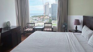 A hotel room with a large bed, a desk with a kettle, two chairs, a nightstand with a lamp, and a view of the city and ocean. - Dragon Sea Hotel (Da Nang)