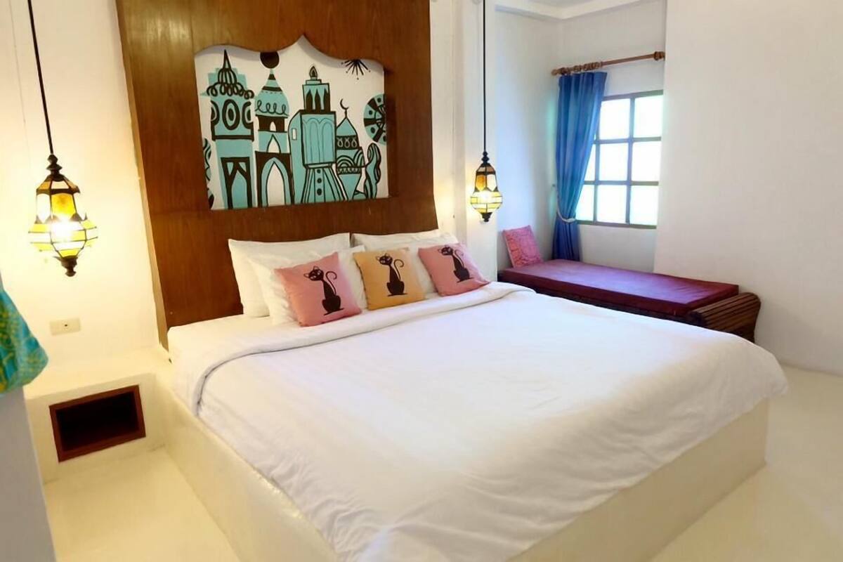 Une chambre d’hôtel avec un grand lit, une tête de lit ornée d’une fresque représentant un paysage urbain, deux lanternes fixées au mur, une fenêtre avec des rideaux bleus et un banc placé près de la fenêtre.