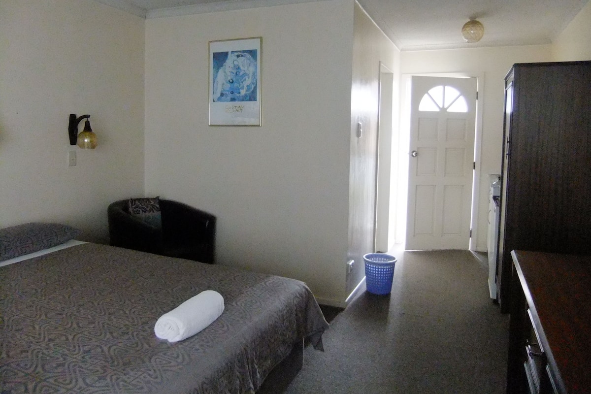 A hotel room with a bed, a desk, a chair, a wall-mounted lamp, a framed picture, and a door leading to another room.
