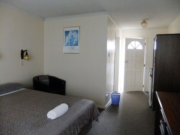 A hotel room with a bed, a desk, a chair, a wall-mounted lamp, a framed picture, and a door leading to another room. - Abbeycourt Motel (Lower Hutt)