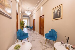Un corridoio d'albergo con pavimento in marmo, poltrone rivestite di blu, un lampadario e opere d'arte incorniciate.
