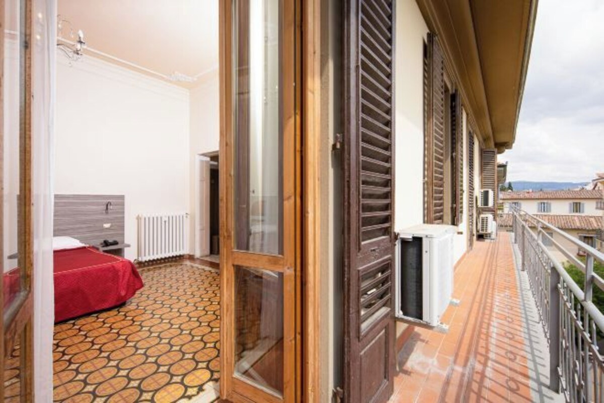 A balcony with a patterned floor, a bed with a red cover, and a view of the cityscape.