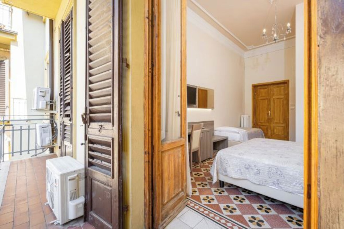 A room with a bed, a desk, and a chair. There is a window with blinds, a door, and a radiator.