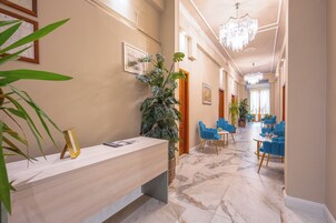 Una hall d'albergo con una reception, sedie blu, lampadari e opere d'arte incorniciate alle pareti.