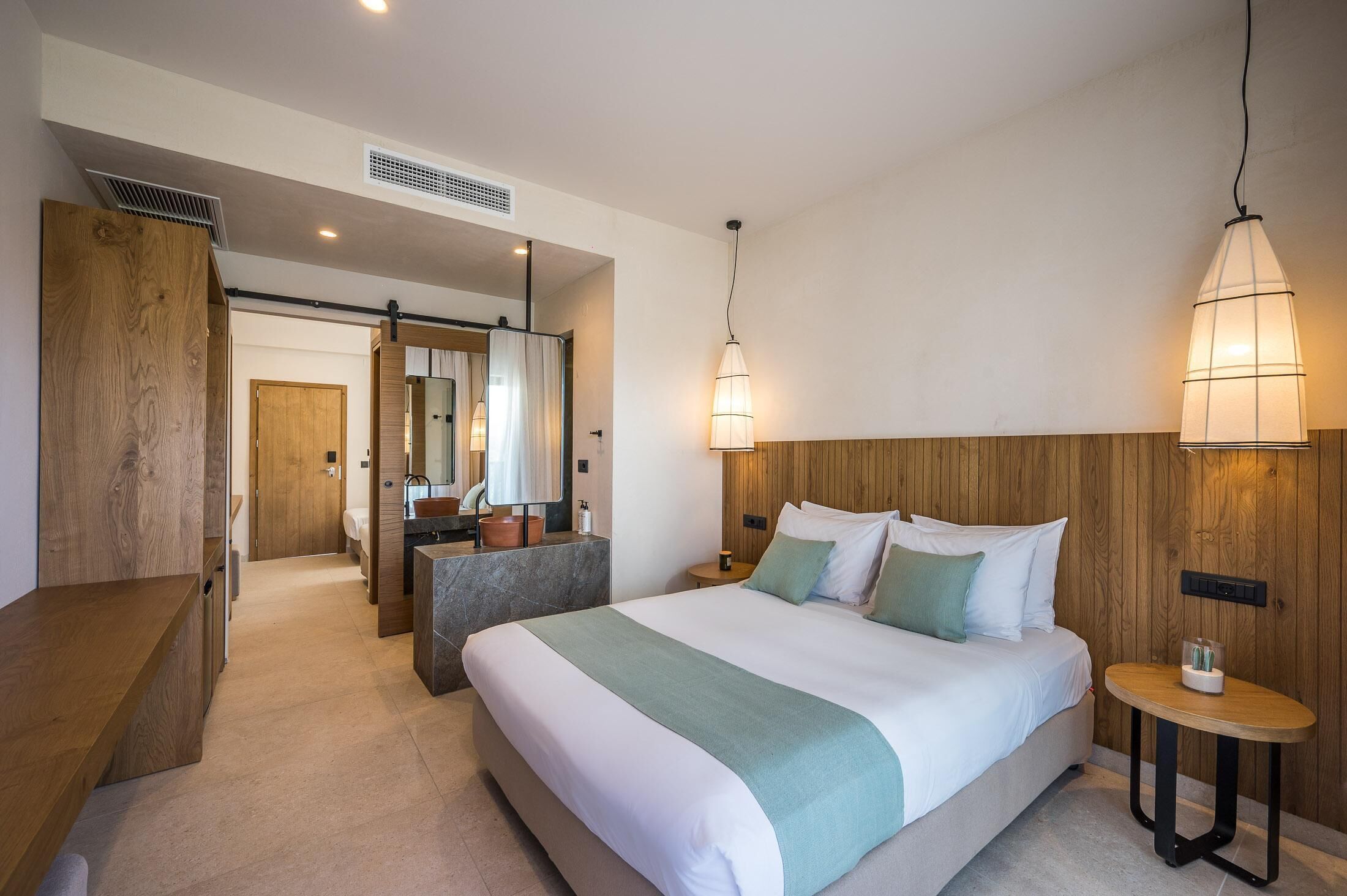 A modern hotel room with a large bed, bedside tables, a small round table, and a wooden headboard.