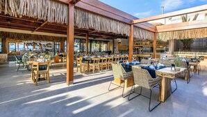 An outdoor dining area with wooden tables and chairs, covered by a wooden pergola with thatch roofing. - Stella Village Seaside Hotel - All inclusive (Hersonissos)