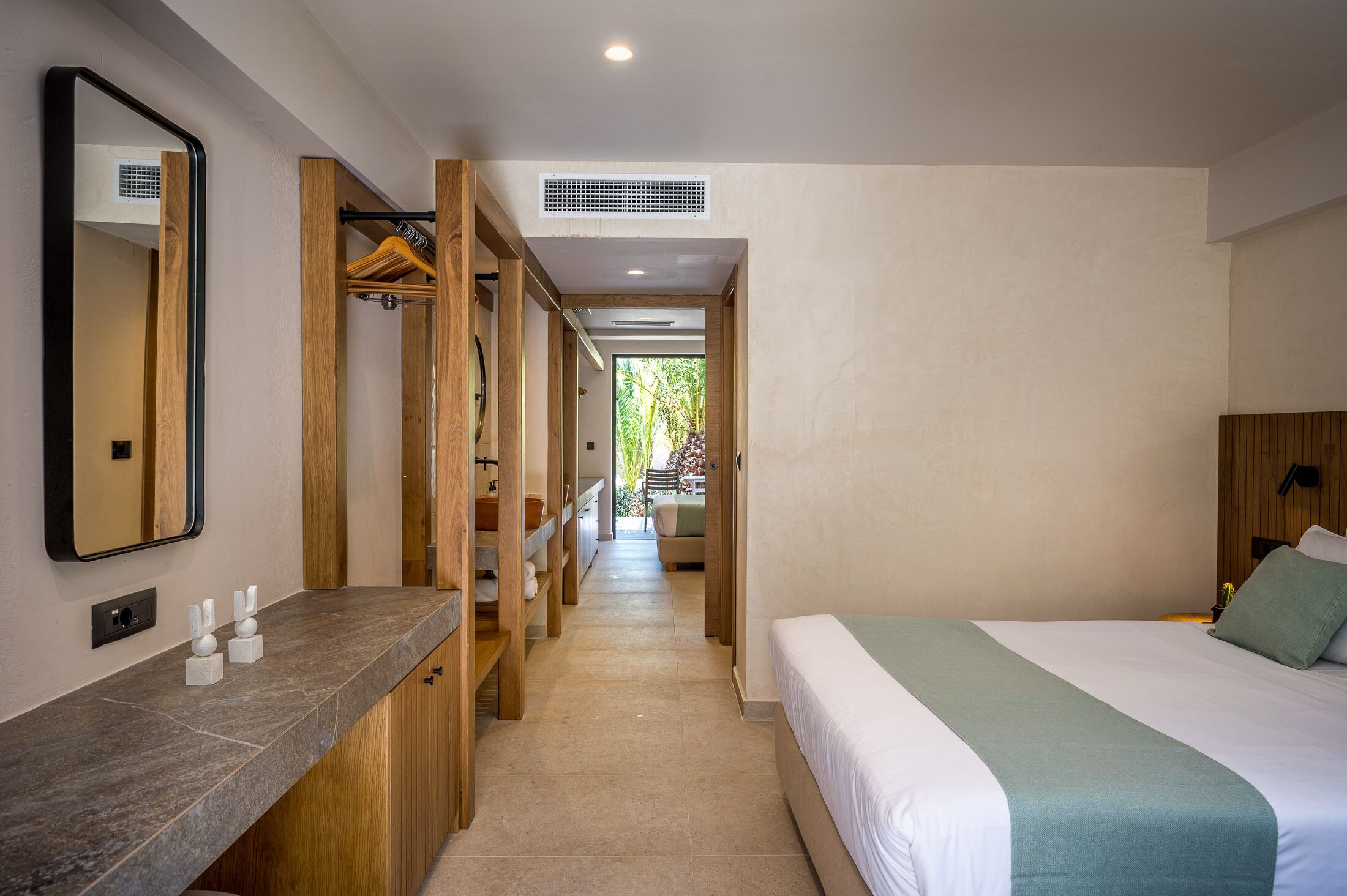 Habitación de hotel con cama, armario de madera, espejo y zona de baño con lavamanos y toallero.