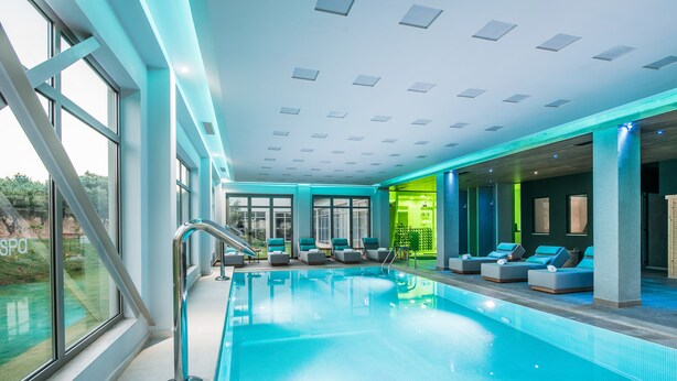 A modern indoor swimming pool with clear glass walls, a blue pool, and lounge chairs. - Stella Village Seaside Hotel - All inclusive (Hersonissos)
