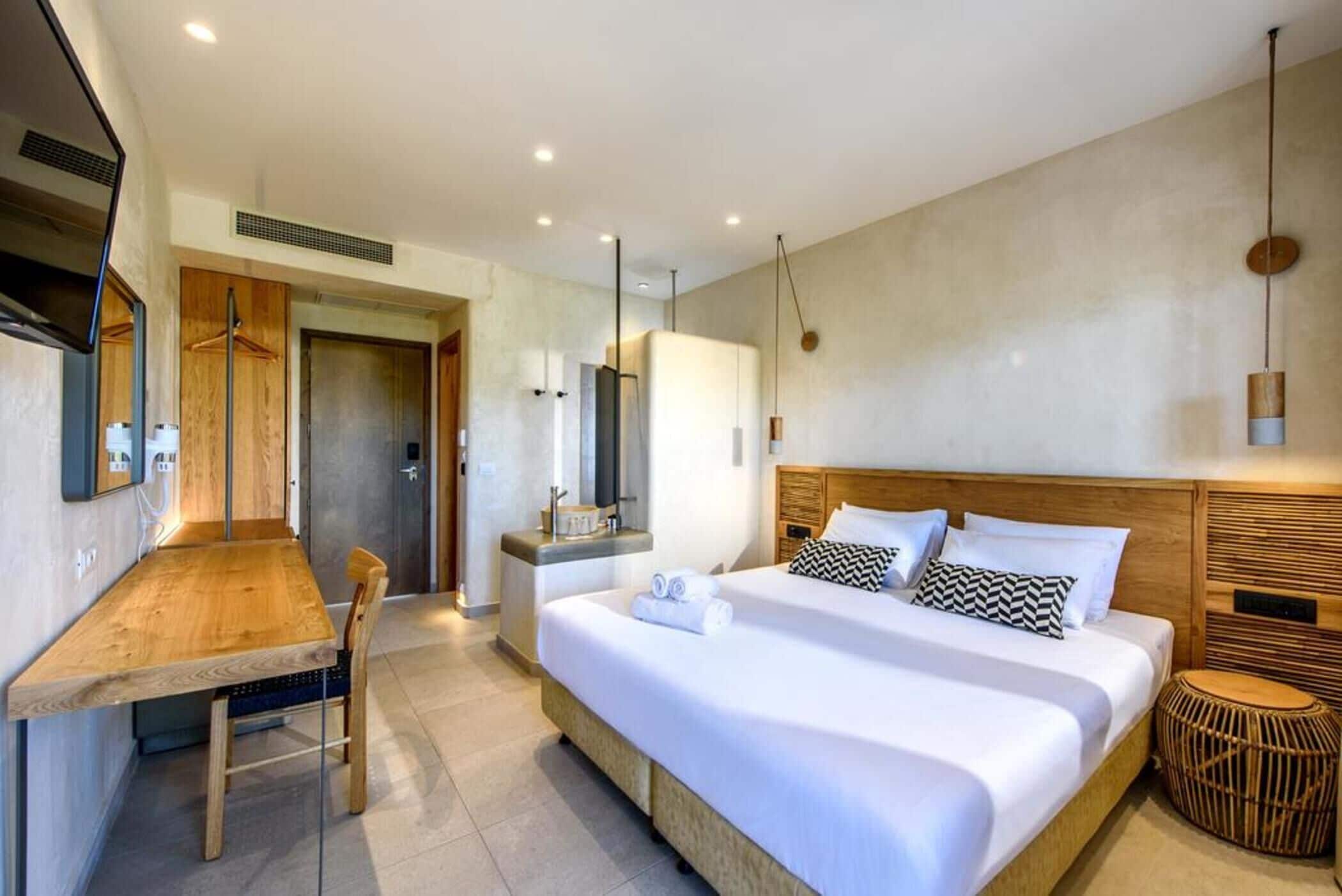 Quarto de hotel moderno com escrivaninha de madeira, cama grande com roupa de cama branca, televisão e mesa redonda pequena.