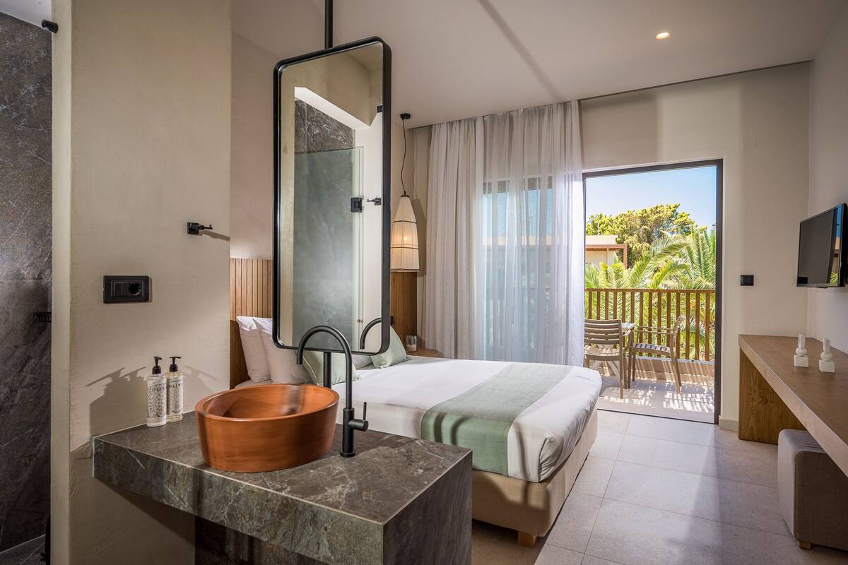 Een moderne hotelkamer met een groot bed, een badkamer met een marmeren wastafel en een balkon met uitzicht op groen.
