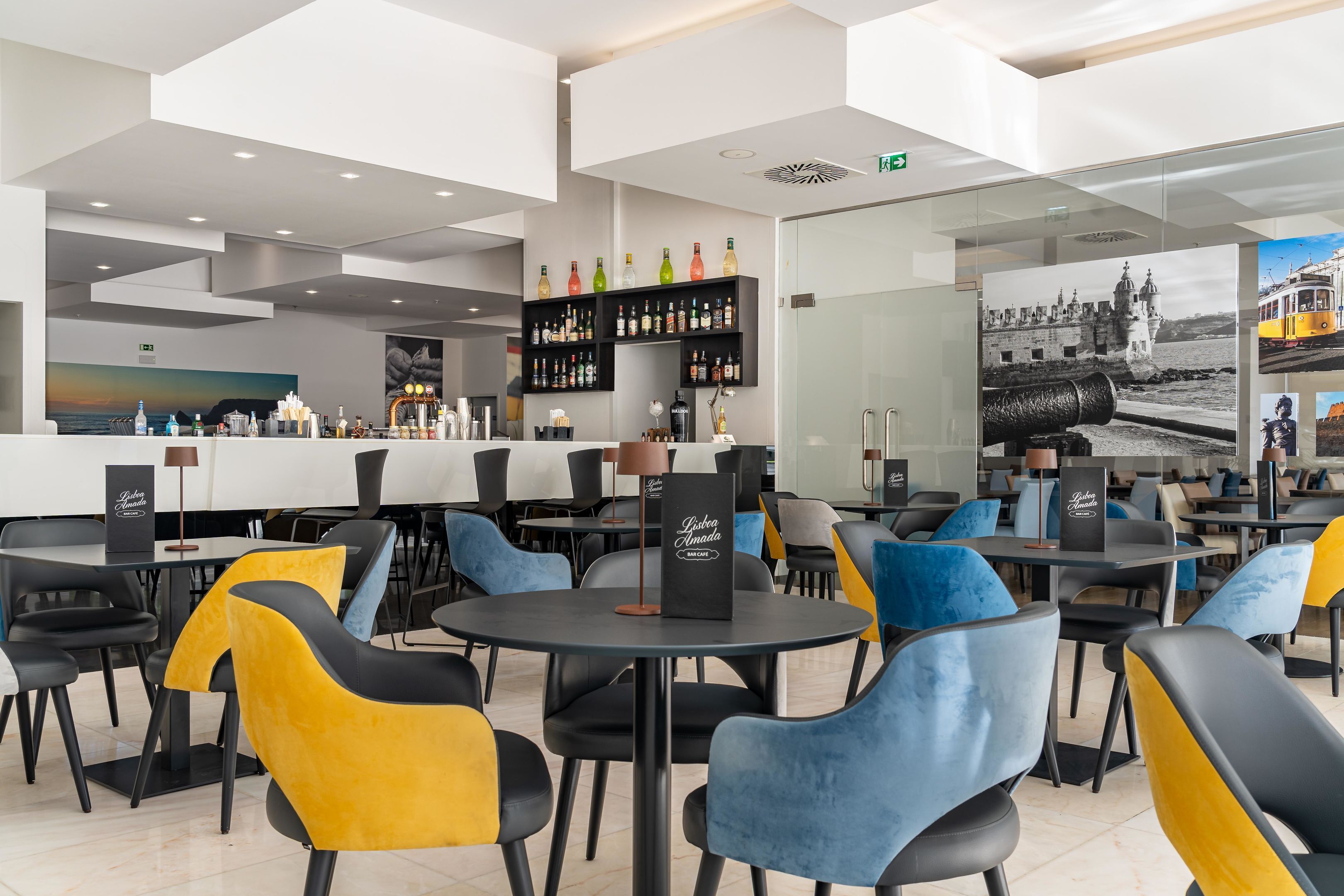 Un restaurante moderno con mesas y sillas redondas, una zona de bar con estantes llenos de botellas y un mural grande que representa una escena costera.