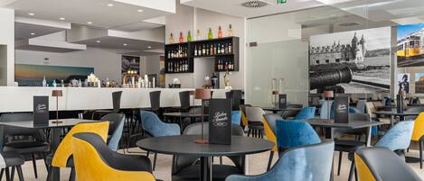 Un restaurante moderno con mesas y sillas redondas, una zona de bar con estantes llenos de botellas y un mural grande que representa una escena costera.