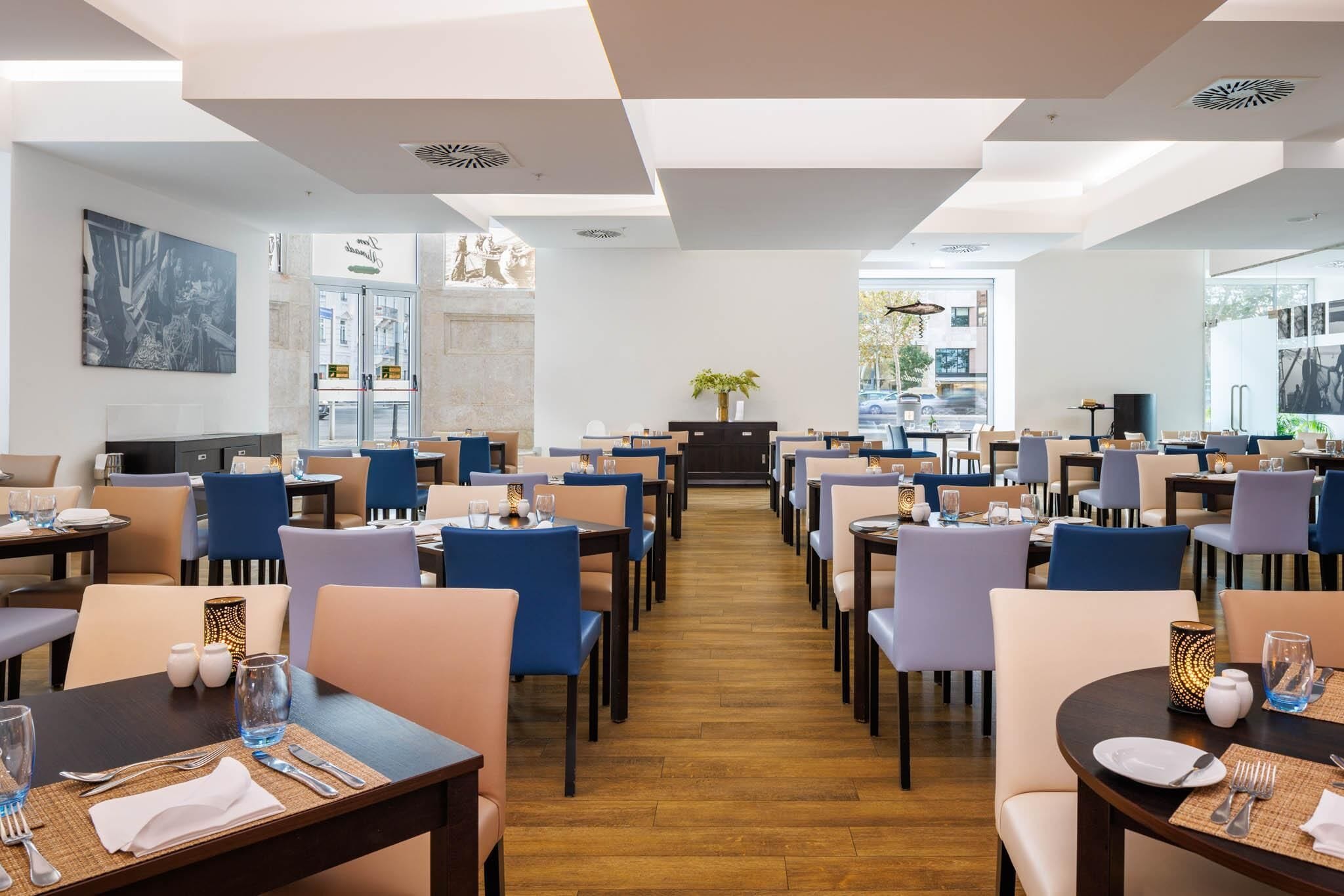 Un restaurant spacieux, doté de nombreuses tables et chaises, d’une plante décorative au centre et offrant une vue sur la rue.