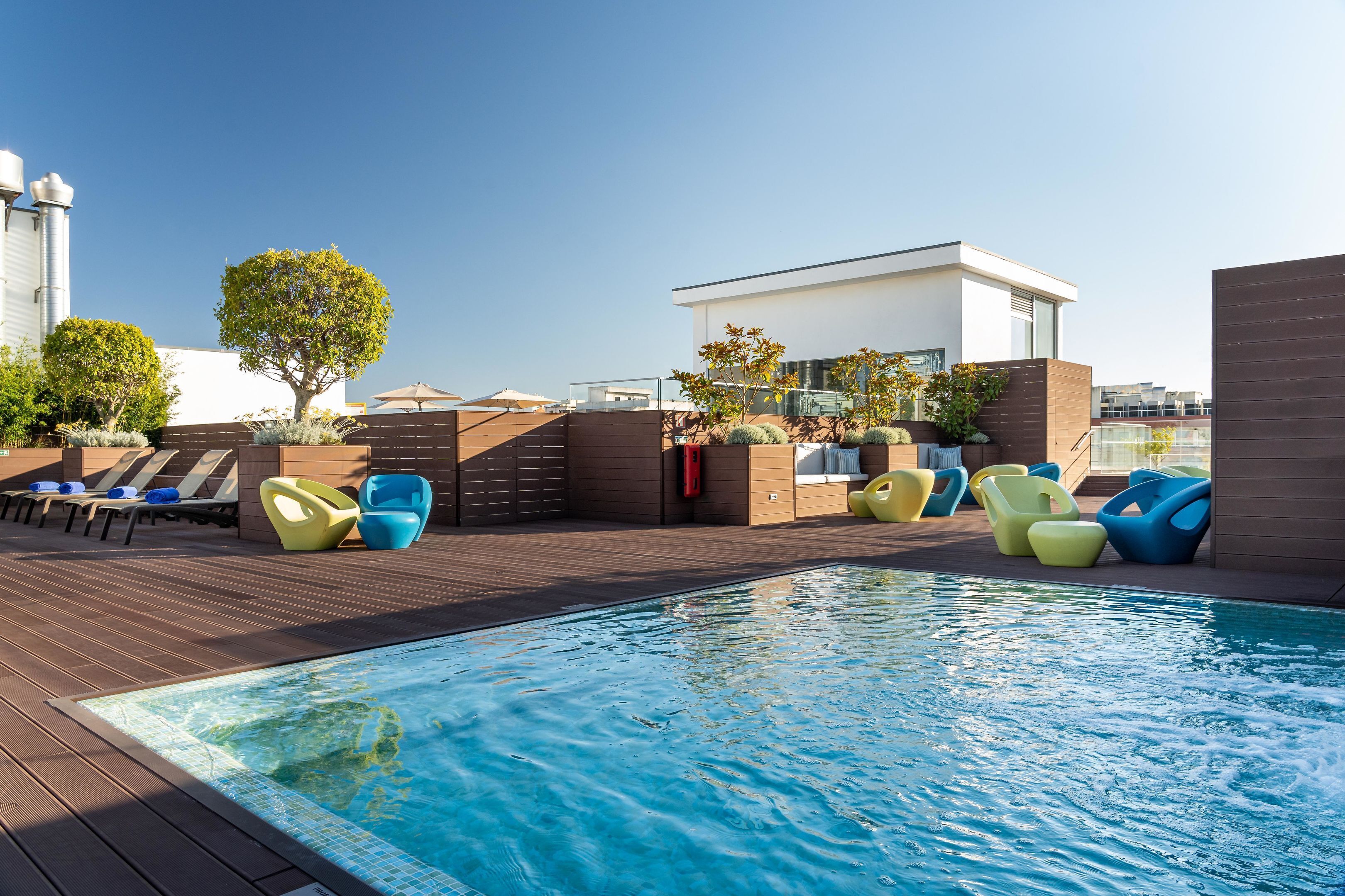 Un espace piscine sur le toit, avec une terrasse en bois, des chaises longues colorées et un bâtiment moderne en arrière-plan.