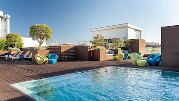 Un espace piscine sur le toit, avec une terrasse en bois, des chaises longues colorées et un bâtiment moderne en arrière-plan.