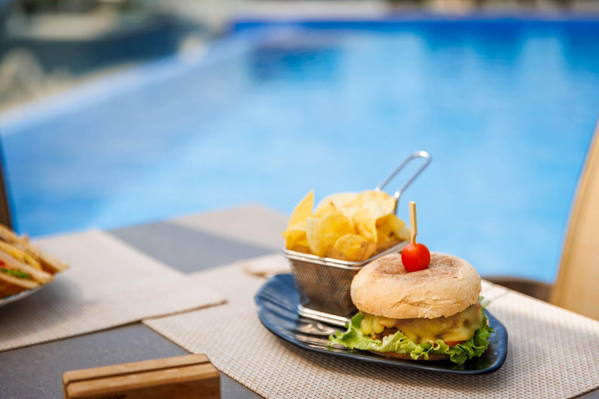 Une table à manger avec une assiette de nourriture, un panier de chips et une piscine en arrière-plan.