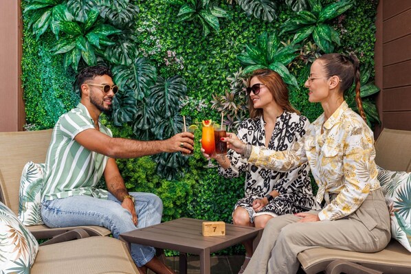 Trois personnes sont assises sur un canapé, avec un mur couvert de plantes vertes derrière elles, et elles tiennent toutes un verre à la main.