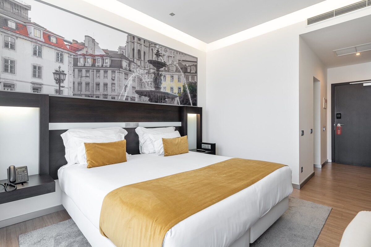 A modern hotel room with a large bed, a headboard with a cityscape mural, a bedside table with a phone, and a door with a handle.