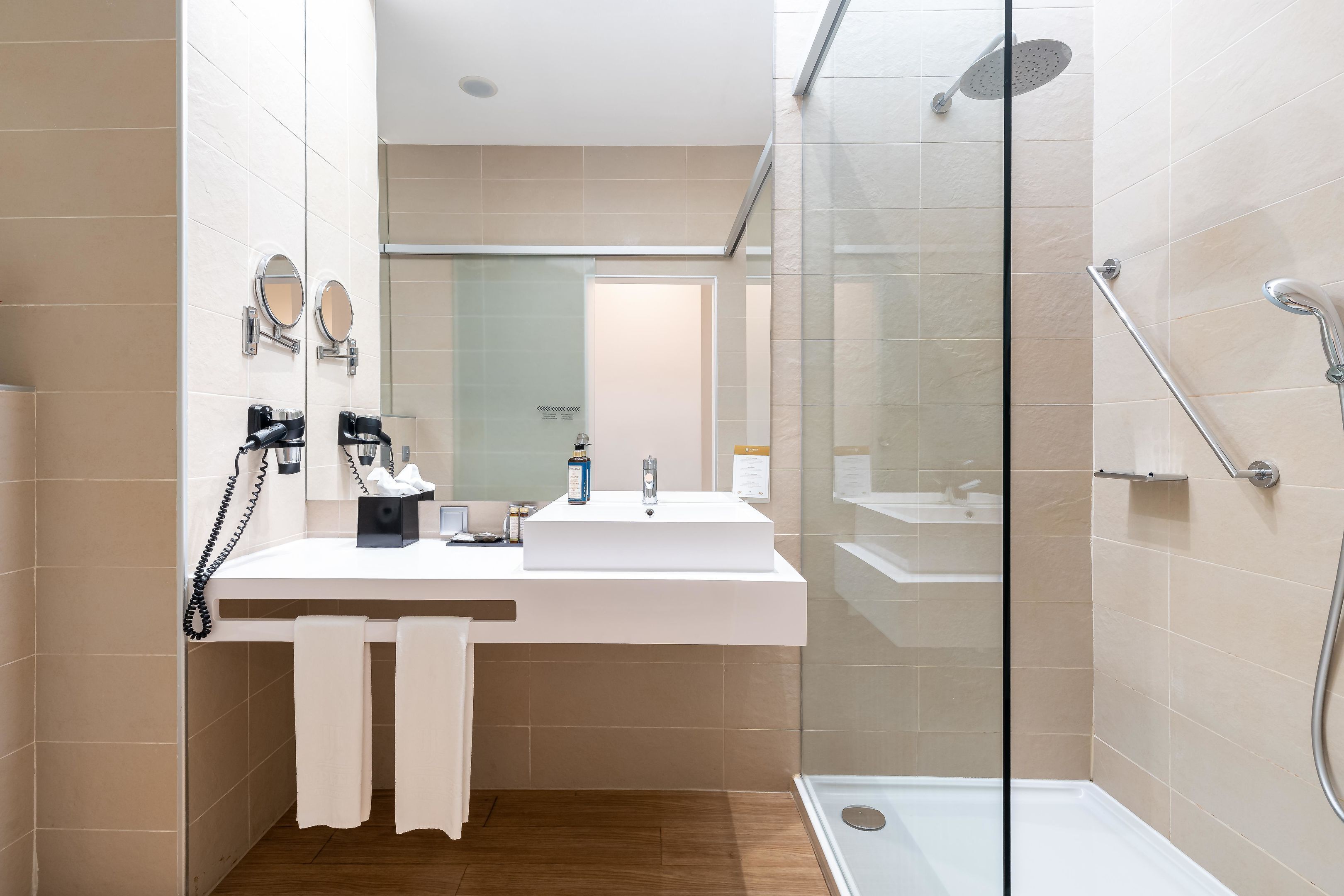 A modern bathroom with a glass-enclosed shower, a white sink with a mirror, and a wooden floor.
