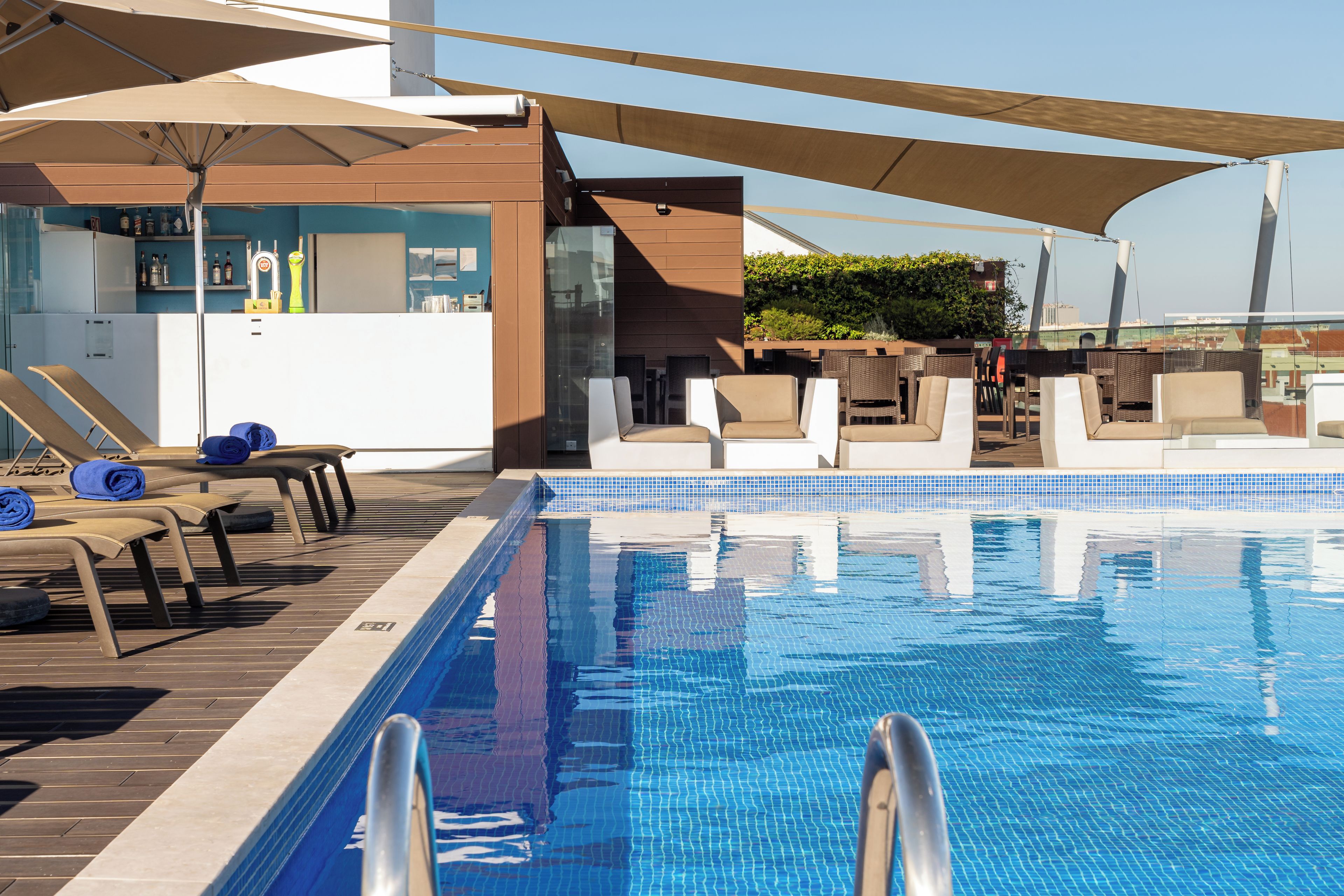 Zona junto a la piscina con sillones y sombrillas, una zona de bar con botellas y vistas a la ciudad.