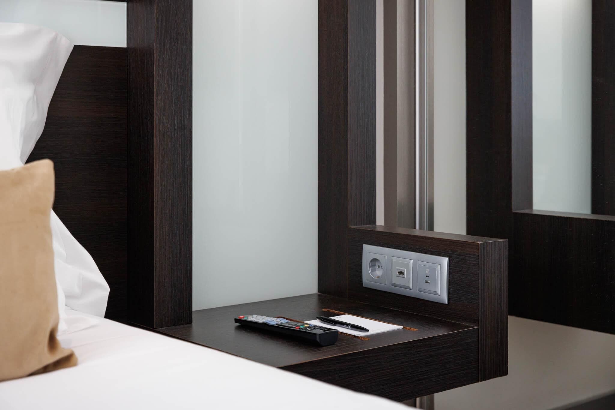 A hotel room with a bed, a nightstand with a remote control, and a wall-mounted power outlet.