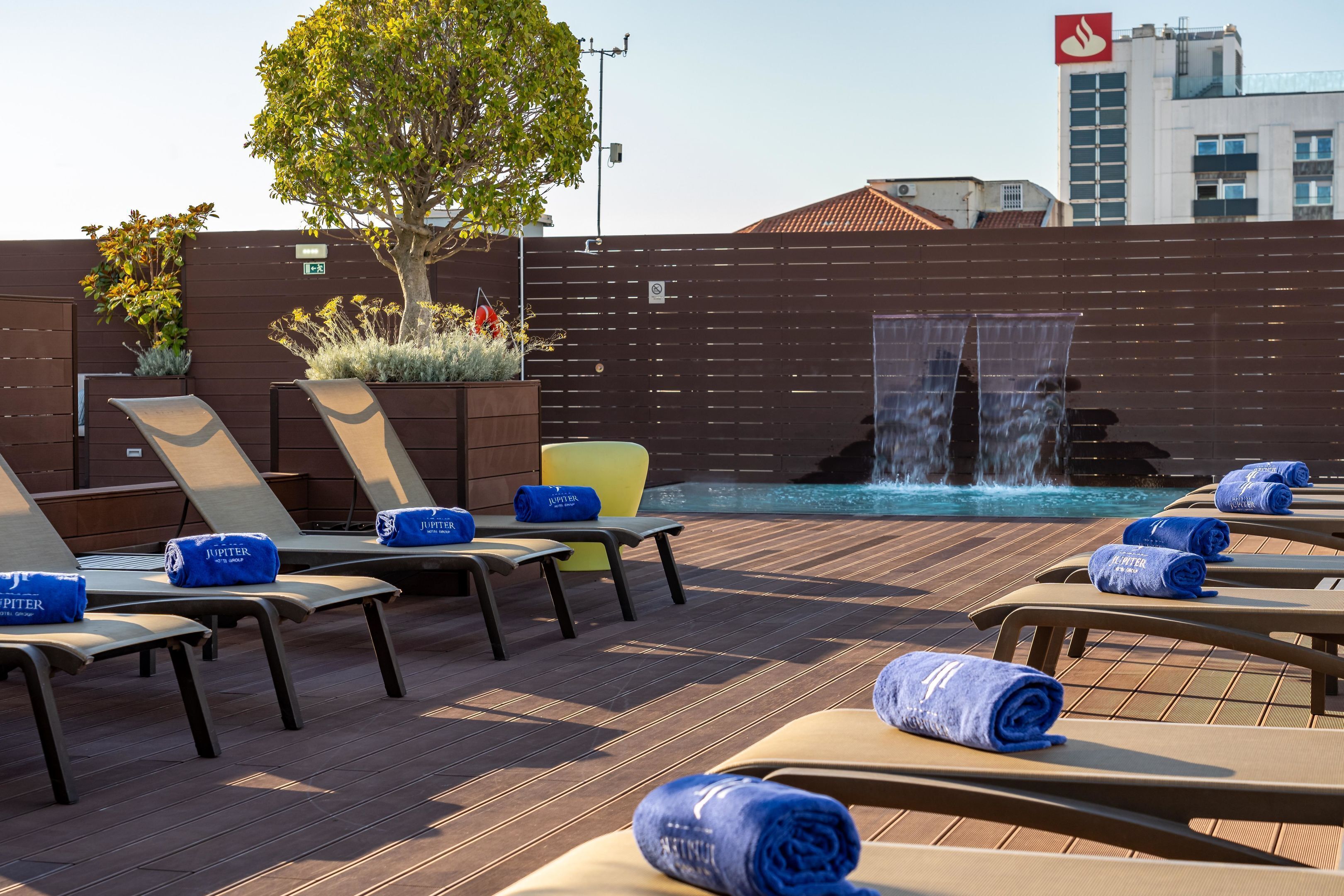A rooftop pool area with sun loungers, a waterfall feature, and a view of a cityscape.