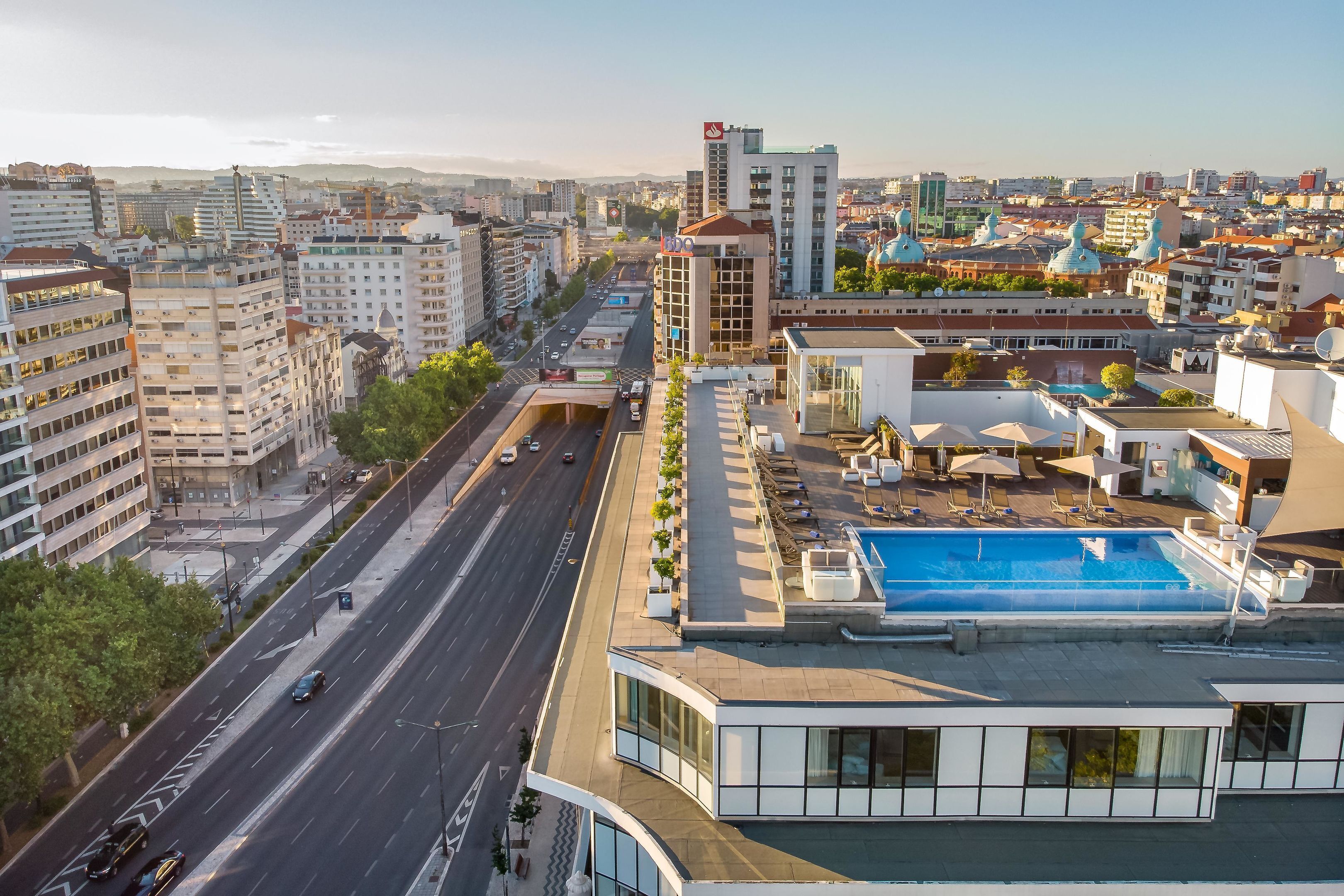 A modern building with a rooftop pool, a spacious road, and surrounding urban buildings.
