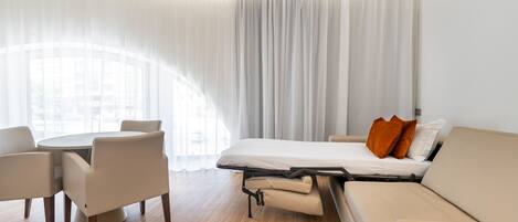 Una habitación de hotel moderna con sofá cama tipo futón, una mesa de comedor con sillas y un ventanal con cortinas traslúcidas.