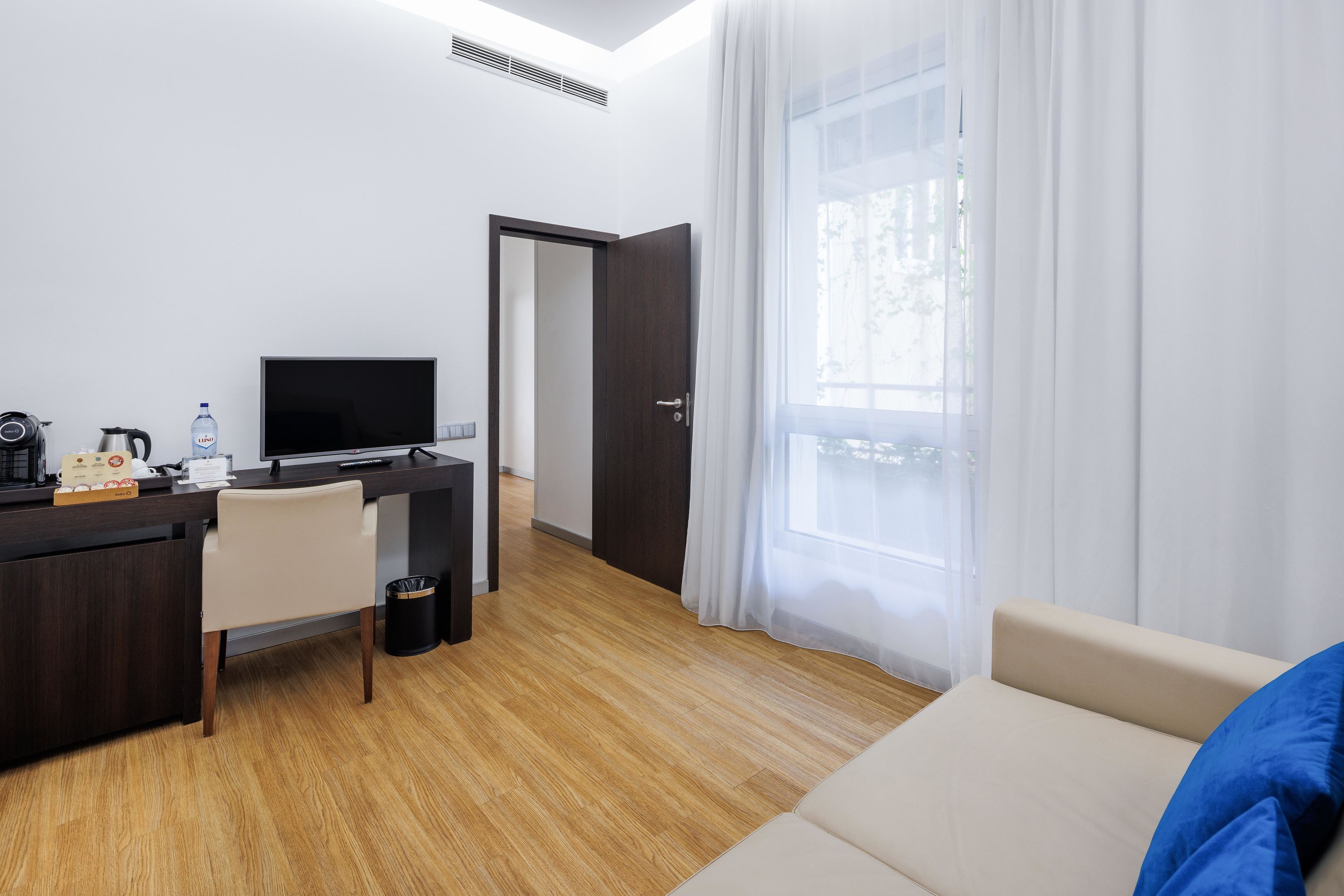Una habitación de hotel con piso de madera, un escritorio con computadora, una silla, una cama con almohada azul y una ventana con cortinas translúcidas.