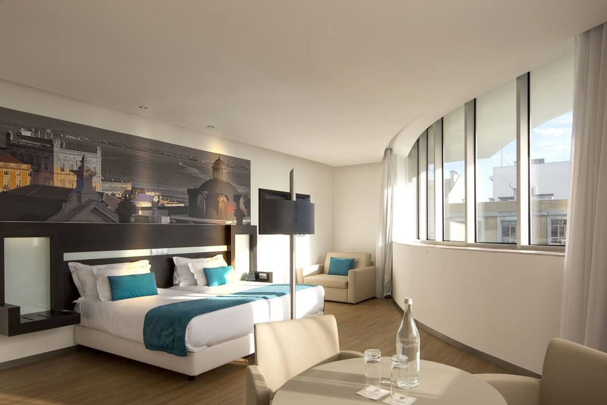 Una habitación de hotel moderna con una cama grande, un espacio de descanso, una mesa con vasos y una botella, y un cuadro con una vista urbana en la pared.