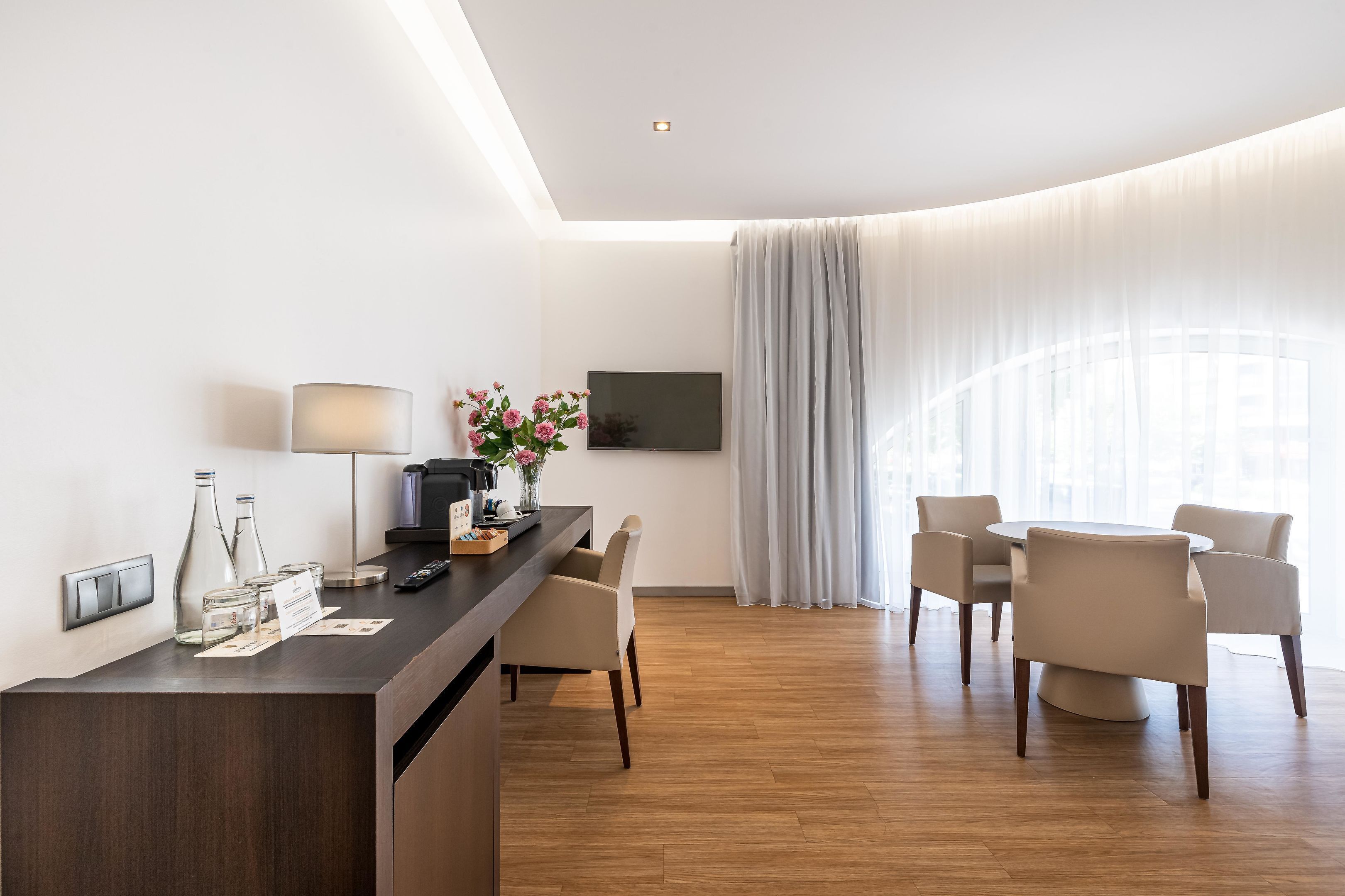 Een moderne hotelkamer met een houten vloer, een ronde eettafel met vier stoelen, een aan de muur bevestigde televisie en een groot raam met dunne gordijnen.