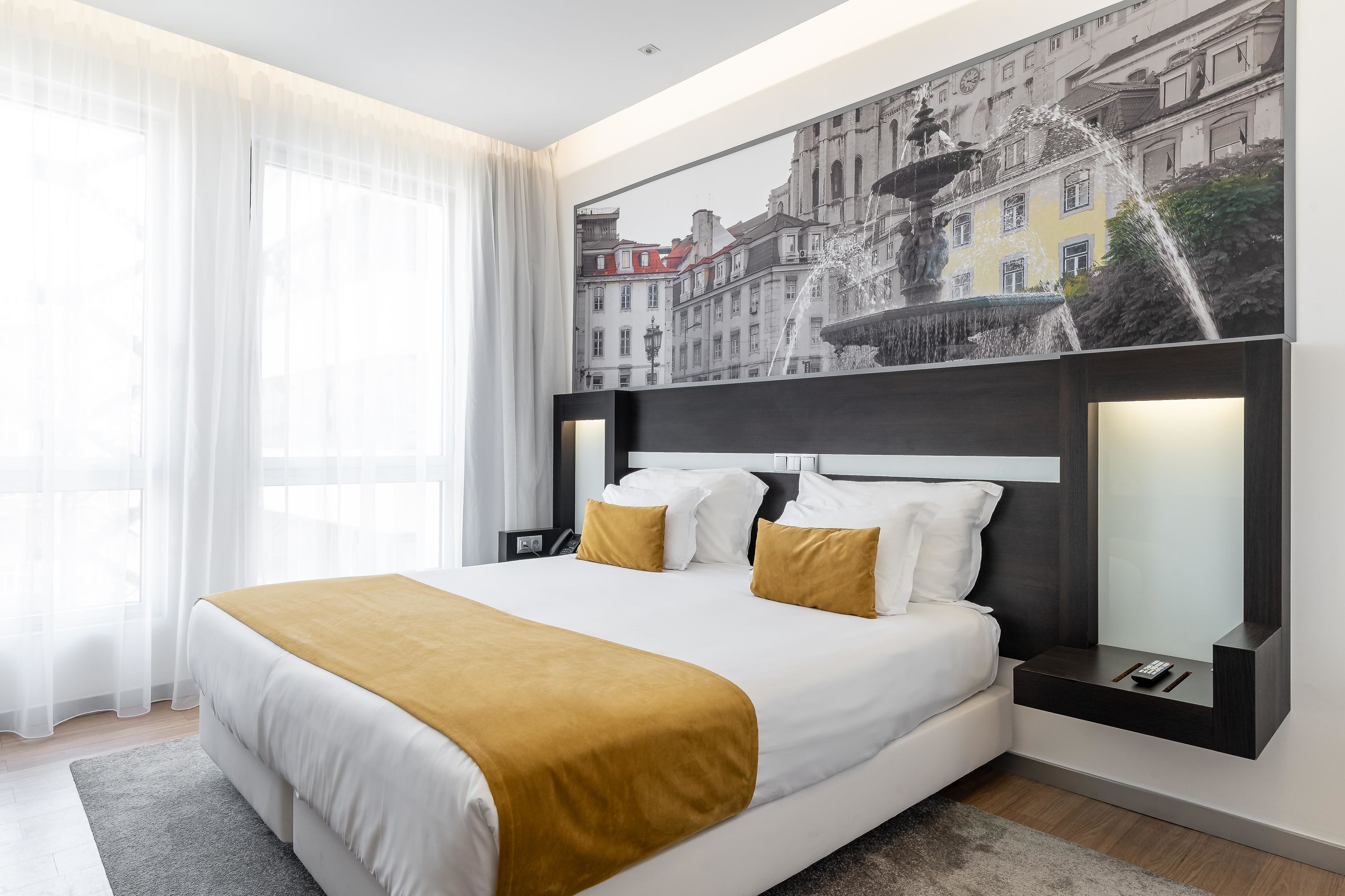 Une chambre d’hôtel moderne avec un grand lit, une table de chevet, un miroir et une œuvre d’art représentant un paysage urbain, accrochée au mur.