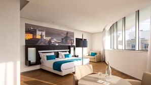 Una habitación de hotel moderna con una cama grande, un sofá, una mesa redonda y un cuadro de un paisaje urbano en la pared.