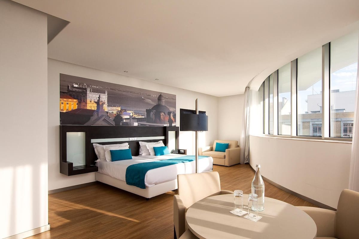 Una habitación de hotel moderna con una cama grande, un sofá, una mesa redonda y un cuadro de un paisaje urbano en la pared.