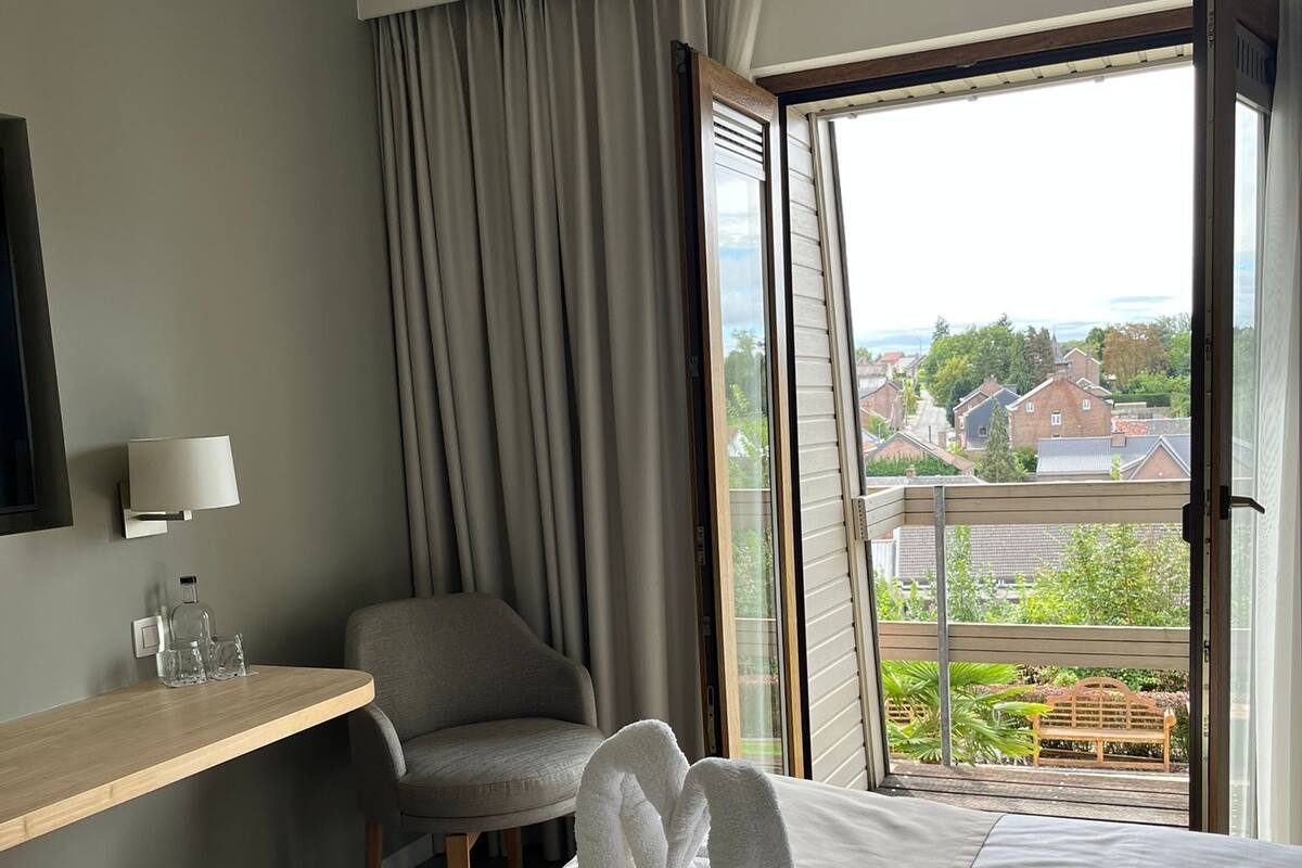 A hotel room with a bed, a chair, a desk with a lamp, and a view of the outside.