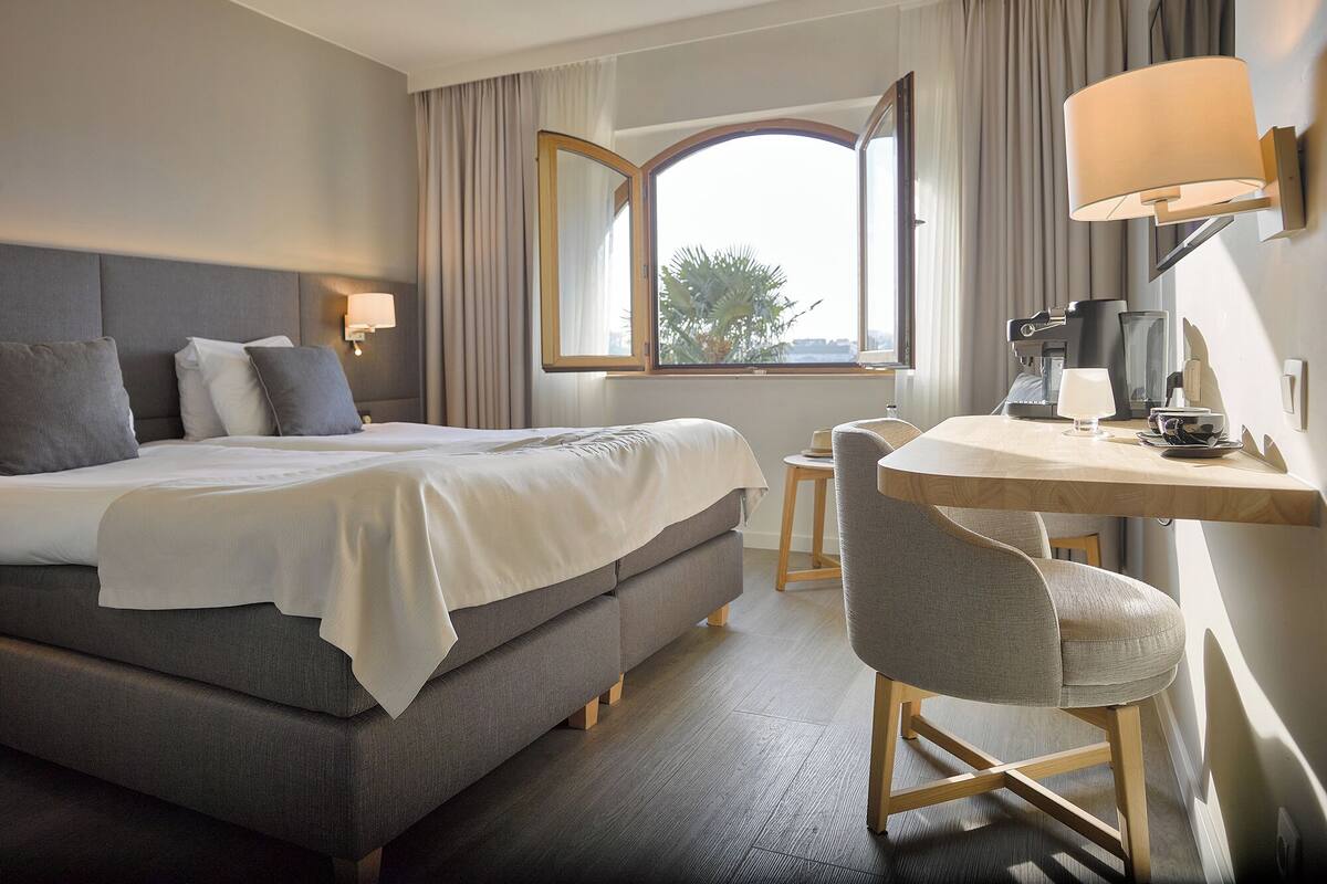A hotel room with a bed, a desk, a chair, a lamp, and a window with curtains.