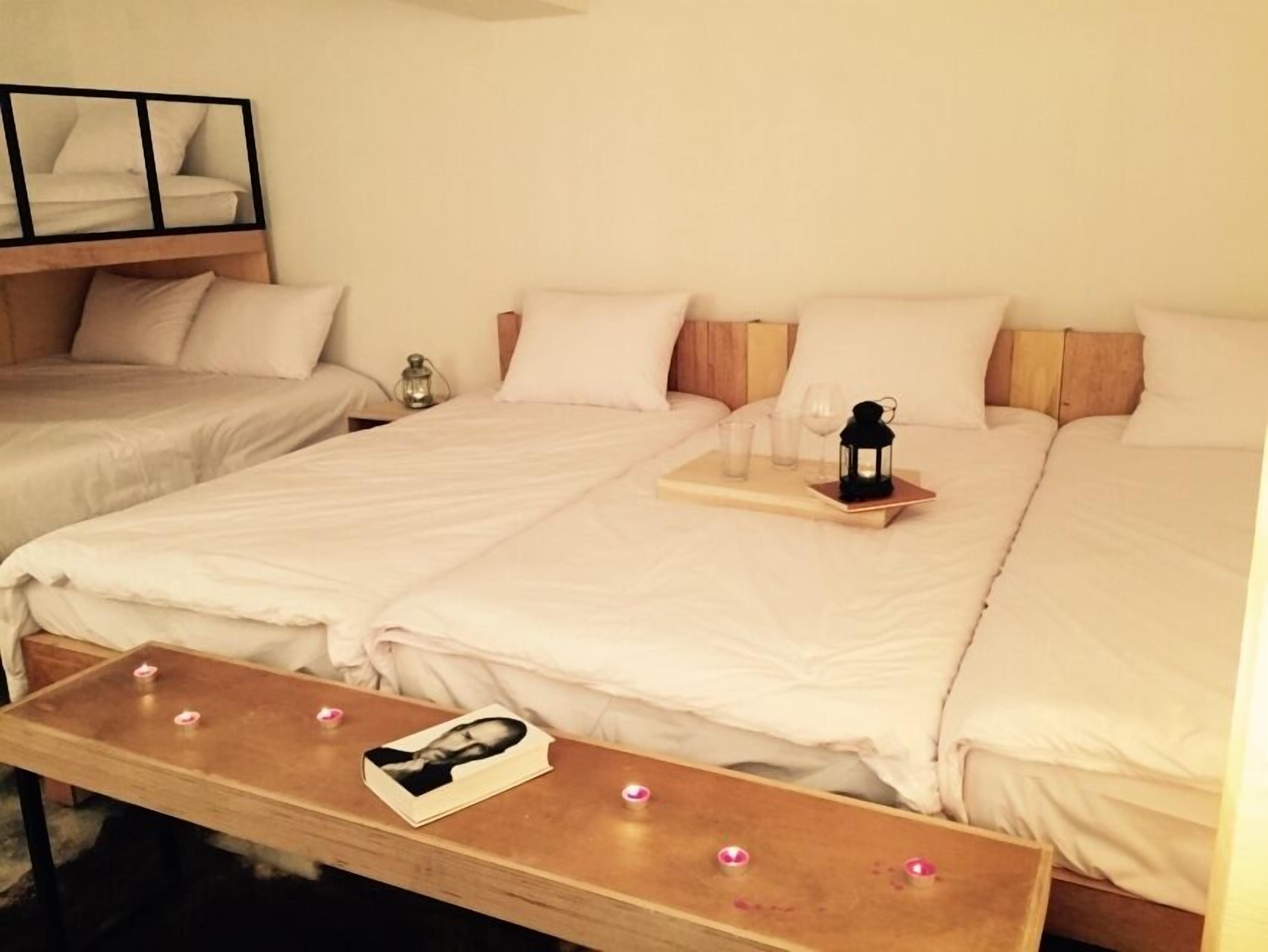 A hotel room with two beds, a bedside table with a photo frame, a small table with glasses and a lantern, and a wall-mounted shelf with framed pictures.