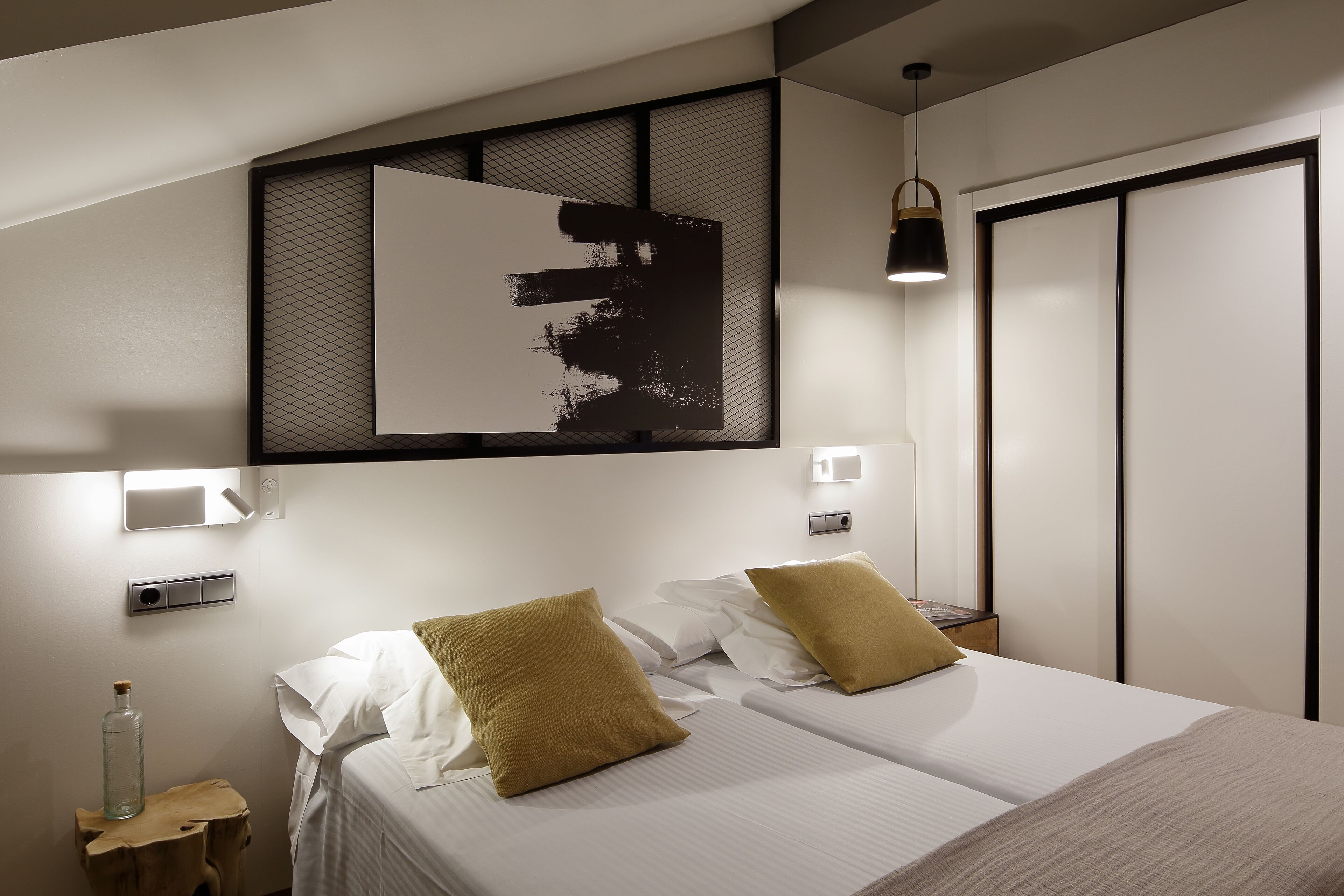 A modern hotel room with a bed, two pillows, a nightstand, a wall-mounted artwork, and a wardrobe.