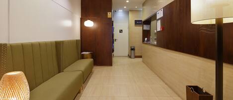 Un hall de hotel con un sofá verde, un cubo de basura, un mostrador de recepción y una lámpara fijada a la pared.
