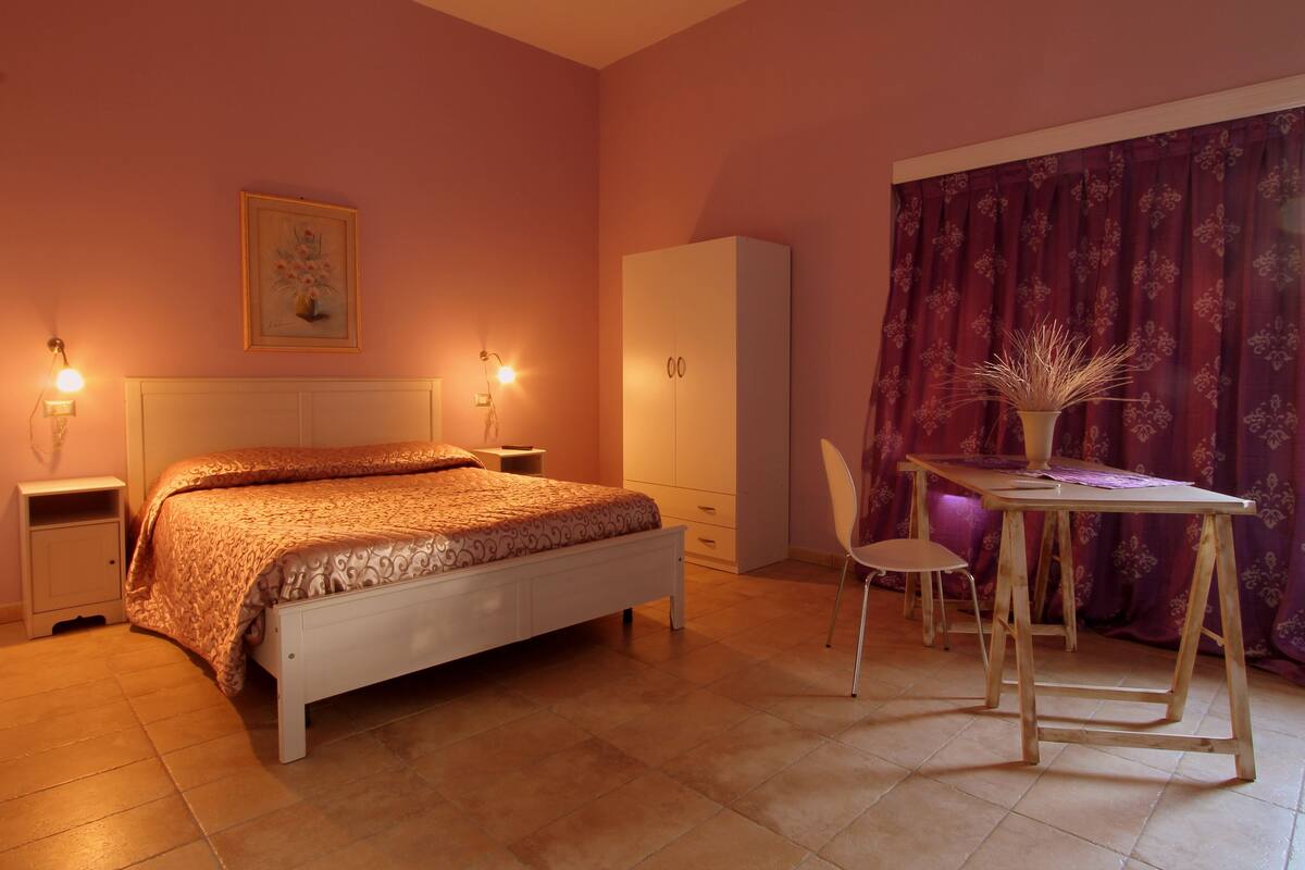 Un dormitorio con cama, mesitas de noche, armario, mesa y sillas, y ventana con cortinas.