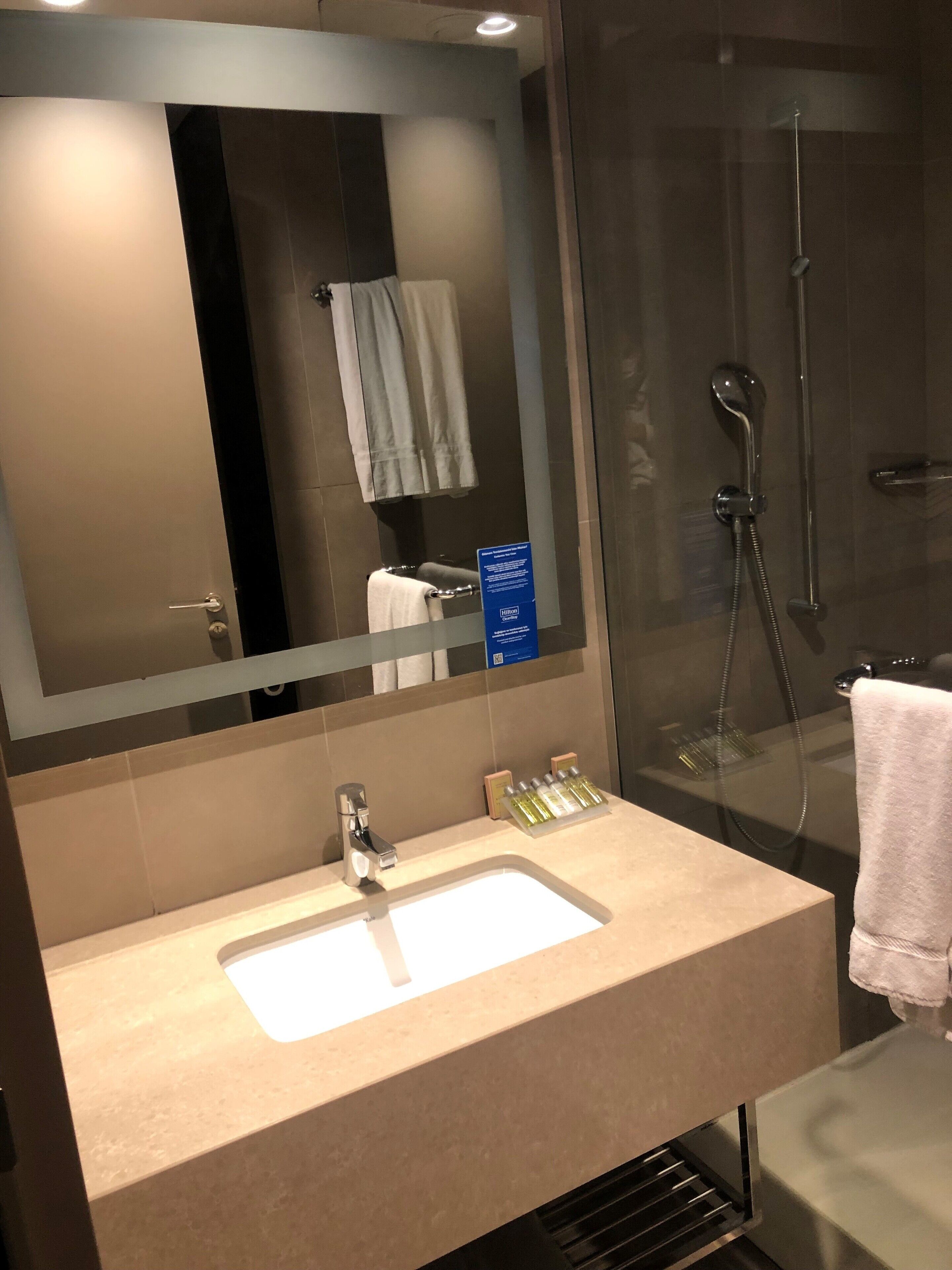 A modern bathroom with a large mirror, a sink with toiletries, a shower, and a towel rack.