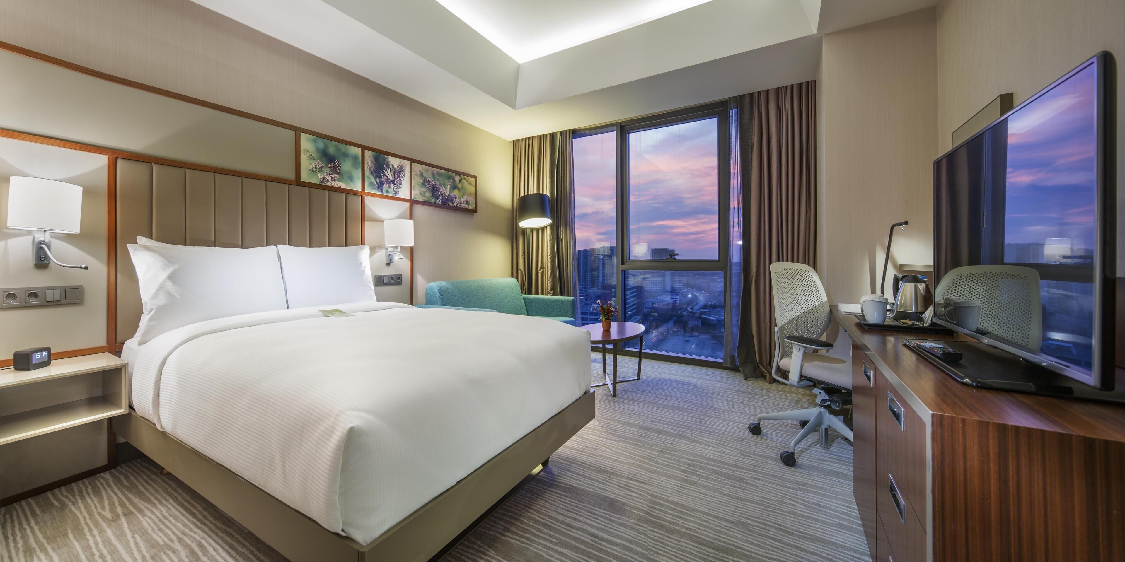 A hotel room with a bed, a desk, a chair, a television, and a large window with a city view.