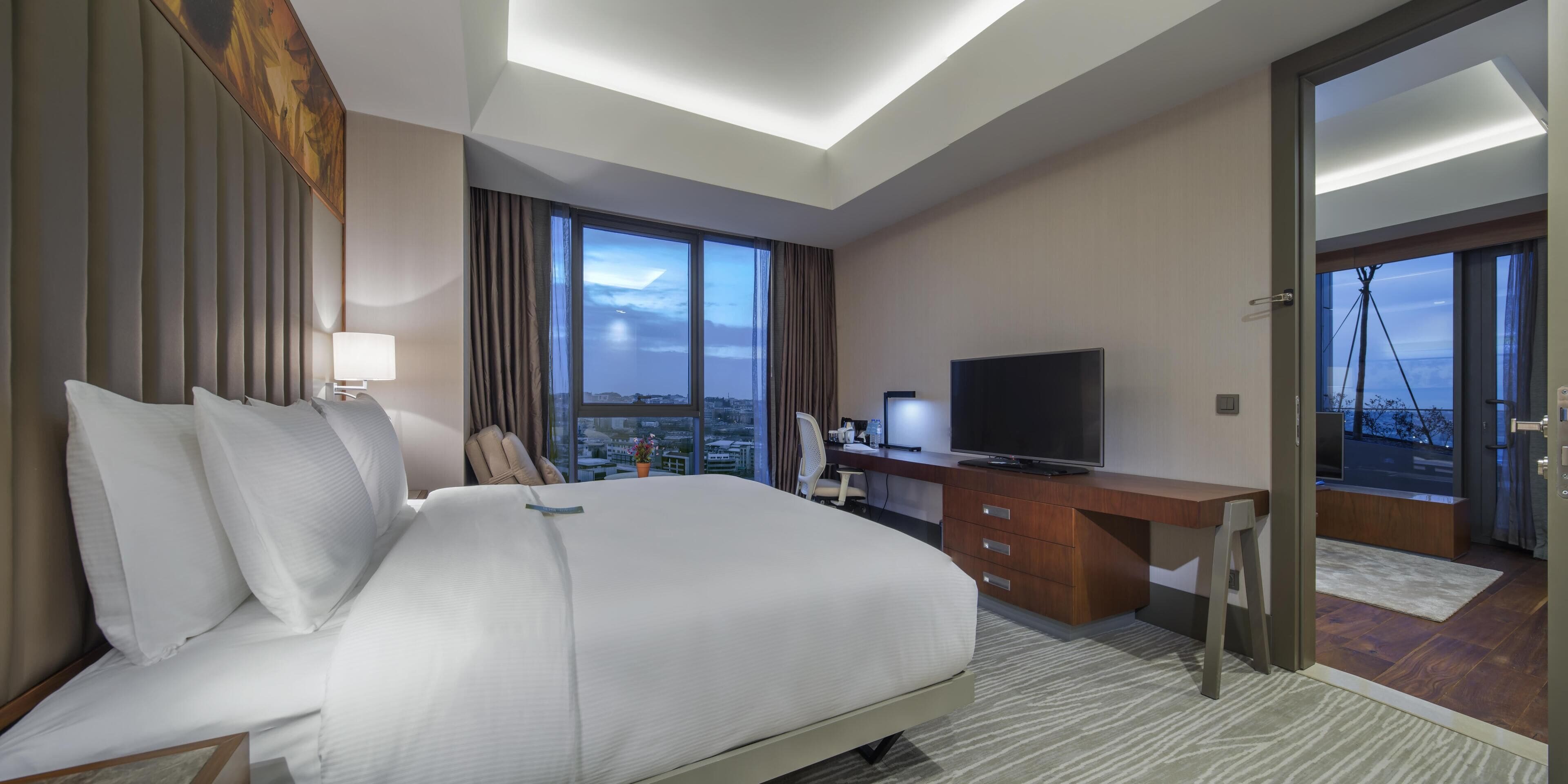 A hotel room with a large bed, a desk with a TV, a chair, and a view of the city through large windows.