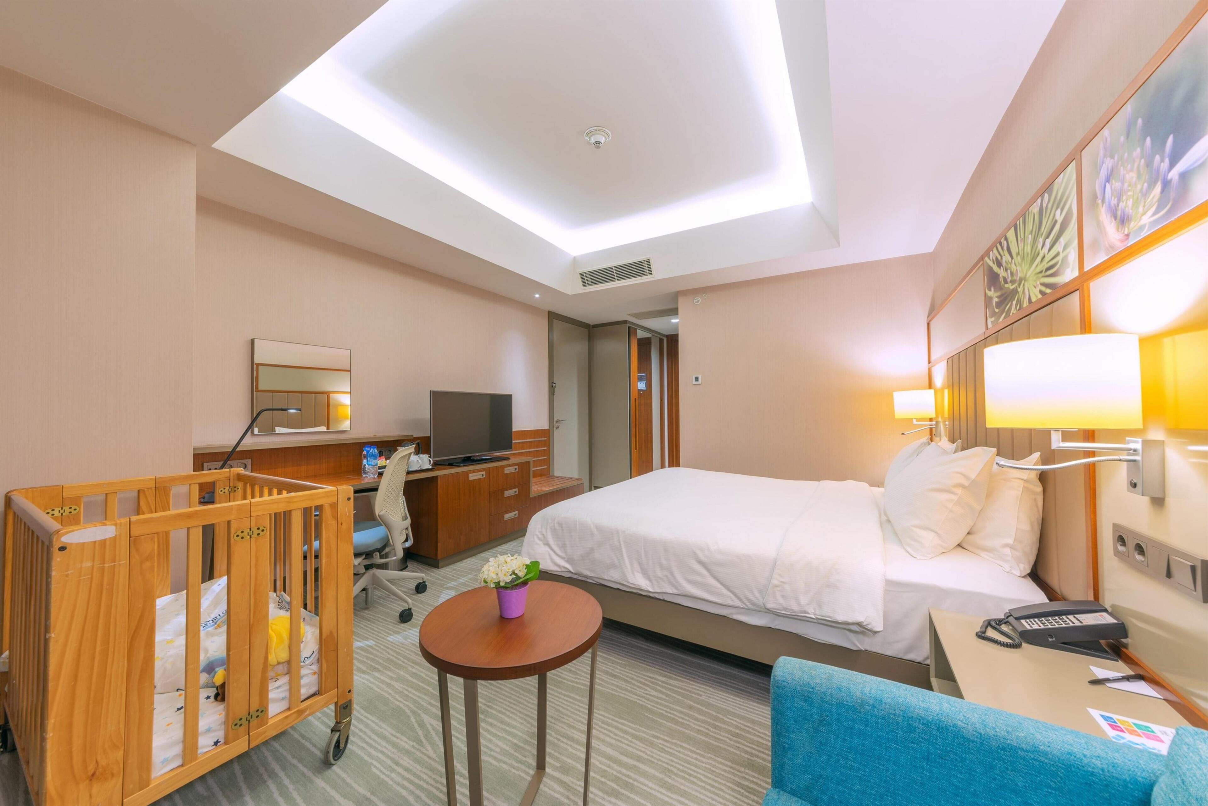 A hotel room with a crib, a bed, a desk, a chair, a television, and a ceiling light.